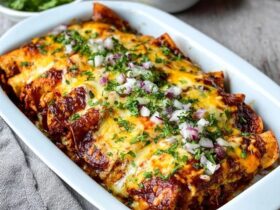 Cheese enchiladas in a casserole dish, rolled corn tortillas covered in deep red enchilada sauce and melted cheese, fresh cilantro, diced red onion on top, sour cream dollops, lime wedges, served in a Mexican ceramic dish