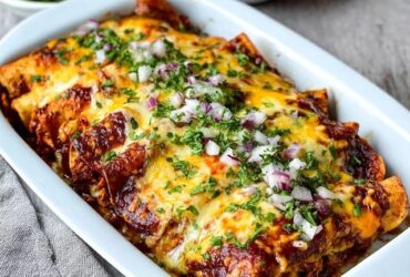 Cheese enchiladas in a casserole dish, rolled corn tortillas covered in deep red enchilada sauce and melted cheese, fresh cilantro, diced red onion on top, sour cream dollops, lime wedges, served in a Mexican ceramic dish
