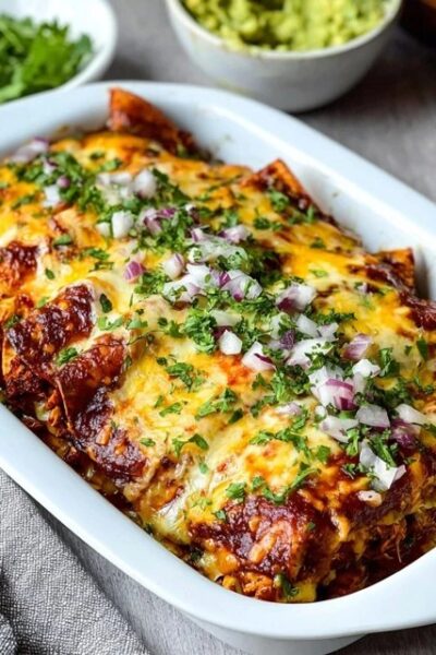 Cheese enchiladas in a casserole dish, rolled corn tortillas covered in deep red enchilada sauce and melted cheese, fresh cilantro, diced red onion on top, sour cream dollops, lime wedges, served in a Mexican ceramic dish
