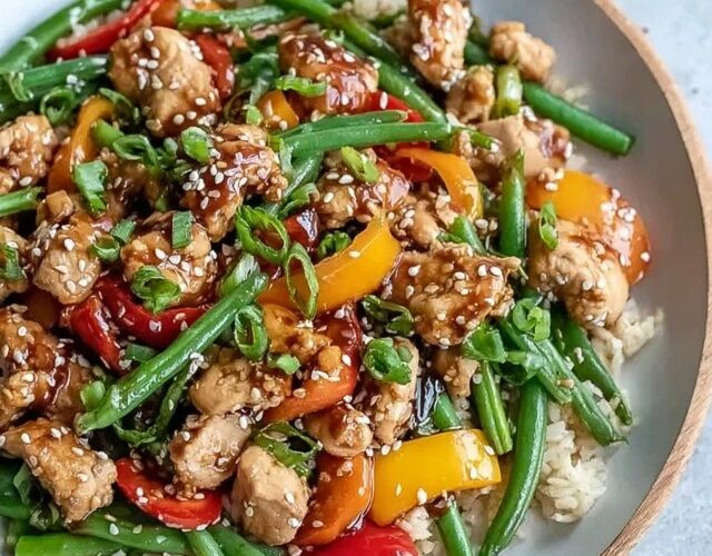 Chicken stir fry with vegetables in a wide black skillet, golden seared chicken pieces, colorful broccoli florets, red and yellow bell peppers, snap peas, sliced carrots, all coated in glossy brown sauce, sesame seeds on top