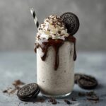 Tall glass of thick Oreo milkshake topped with whipped cream, a whole Oreo on the rim, crushed cookie crumbs, chocolate drizzle down the inside of the glass, striped straw