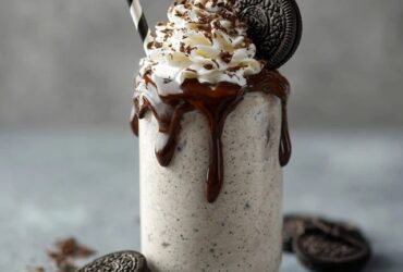 Tall glass of thick Oreo milkshake topped with whipped cream, a whole Oreo on the rim, crushed cookie crumbs, chocolate drizzle down the inside of the glass, striped straw