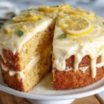 Lemon drizzle cake loaf on a board, golden crumb with a crackly sugar-lemon crust on top, a few slices cut to show the moist tender interior, lemon slices and zest alongside
