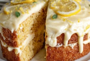 Lemon drizzle cake loaf on a board, golden crumb with a crackly sugar-lemon crust on top, a few slices cut to show the moist tender interior, lemon slices and zest alongside