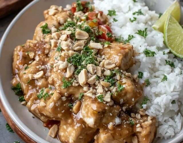 Peanut chicken over fluffy white rice in a bowl, glossy golden-brown peanut sauce coating tender chicken pieces, topped with crushed peanuts, sliced green onion, lime wedge and cilantro