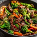 Beef and broccoli stir fry in a wok, glossy dark brown sauce coating tender beef strips and bright green broccoli florets, sesame seeds and sliced green onion on top, served over white rice
