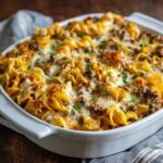Beef noodle casserole in a baking dish, egg noodles in a creamy tomato beef sauce topped with bubbly golden melted cheese, a scoop removed to show the cheesy layers