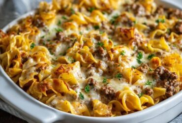 Beef noodle casserole in a baking dish, egg noodles in a creamy tomato beef sauce topped with bubbly golden melted cheese, a scoop removed to show the cheesy layers