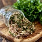 Homemade herb and spice blend in a small glass jar and spilled on a spoon, a rich brown-red seasoning mix with visible dried herbs, paprika and pepper flakes
