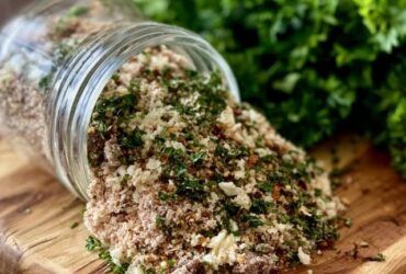 Homemade herb and spice blend in a small glass jar and spilled on a spoon, a rich brown-red seasoning mix with visible dried herbs, paprika and pepper flakes