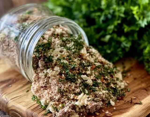 Homemade herb and spice blend in a small glass jar and spilled on a spoon, a rich brown-red seasoning mix with visible dried herbs, paprika and pepper flakes